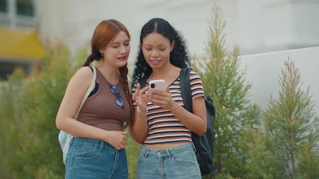 Two young women stand together, sharing a phone. They smile, engaged in conversation. Green plants frame their casual outdoor moment. Perfect for social media or student lifestyle scenes