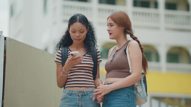Two young women stand together, sharing a phone. They smile, engaged in conversation. Green plants frame their casual outdoor moment. Perfect for social media or student lifestyle scenes