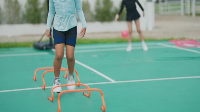 Young girl runs swiftly across green court. She leaps over orange hurdles with focus and joy. Her curly hair flies as she moves with energy. Background shows another child near basketball hoop