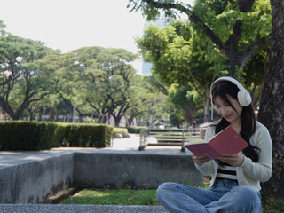 Fototapeta premium Young Thai Student Wearing Headphones Reading a Pink Book in a Park