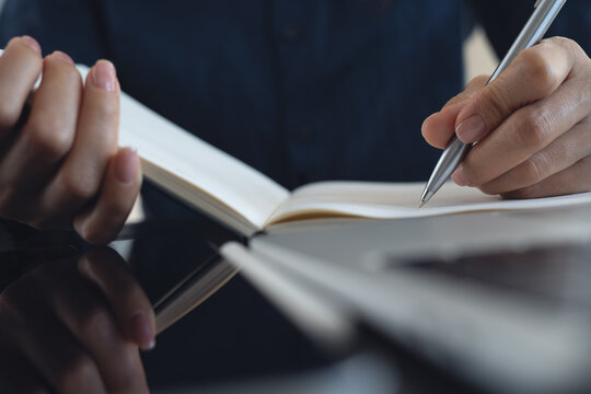 Close up, woman hand with a pen writing in notebook with laptop on office desk, planning and making a note reminder