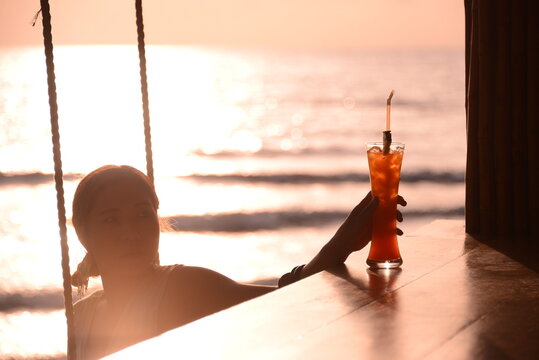 Woman sitting on the swing grabbing a tall glass of cold drink at beach bar during sunset time