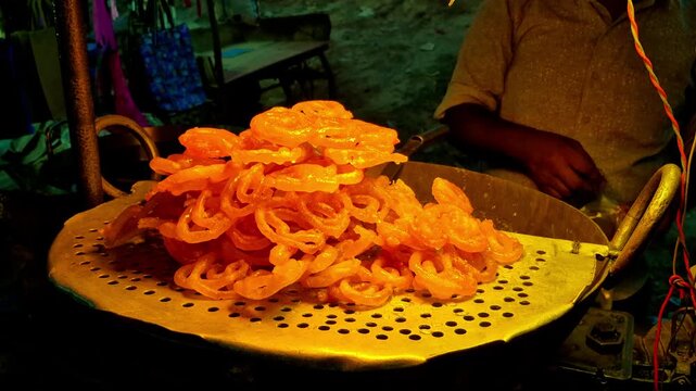 Freshly prepared jalebis form a vibrant mound glistening under warm market lights at the lively evening sweet stall. A street vendor readies newspaper wrapping for customers browsing glowing display.