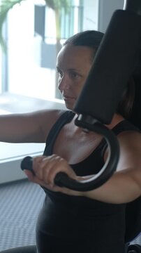 Vertical video. Young athletic woman sitting at a chest press machine in a modern gym, pushing the handles forward with focused concentration during a strength training session