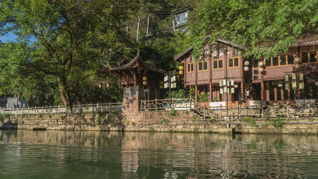Wooden buildings with curved tiled roofs are on the river embankment. Green vegetation. Stone barrier and railings along the riverbed. Reflection on the shiny water. China. Fenghuang Ancient Town
 