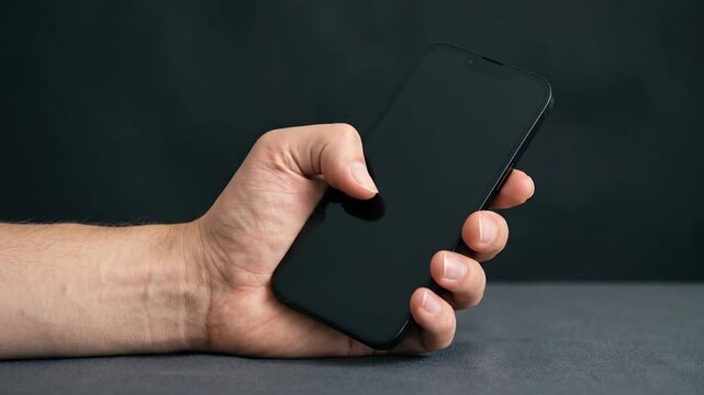 Close-up of man holding and tapping blank smartphone screen against dark background.
