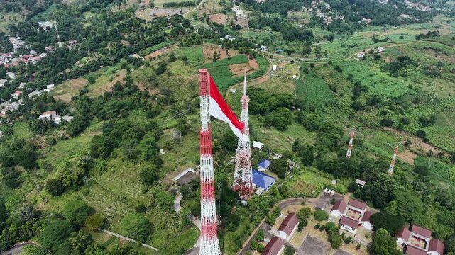 Drone aerial footage of the Indonesian national flag waving on a communication tower above a rural landscape. Patriotic red and white flag symbolizing Indonesia's independence, national pride, freedom