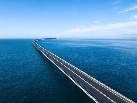 A long curved sea bridge stretches across clear blue ocean under bright sky, with smooth road and supporting piers.