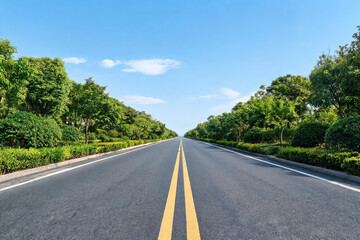 Fototapeta premium A straight empty asphalt road with double yellow lines, flanked by lush green trees and shrubs under a clear blue sky.