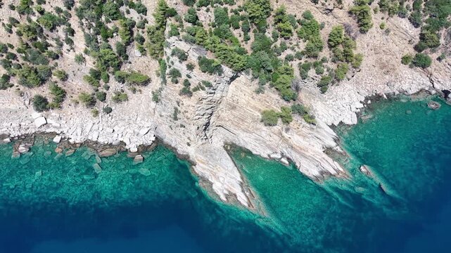 Oblique aerial view moving along a rugged stone coast with transparent turquoise shallows and visible submerged rocks.