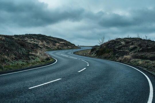 A winding paved road with lane markings cuts through hilly moorland under an overcast, cloudy sky, in a remote rural setting.