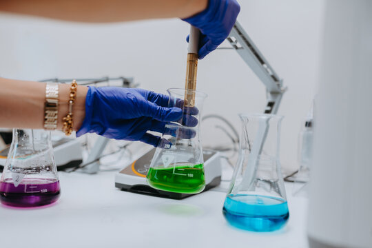 Laboratory Scientist With Gloved Hands Pipetting Colorful Chemical Samples Into Erlenmeyer Flasks For Research