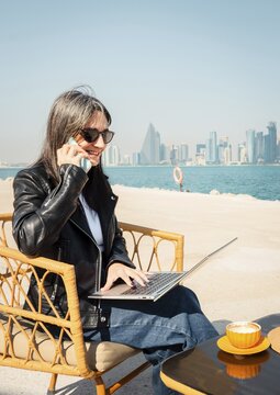 Happy business woman drinking coffee using laptop and smartphone chat. West Bay skyline background digital nomad working remotely in outdoor cafe. Middle East city urban professional. Doha.Qatar
