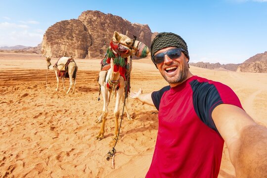 Smiling man traveler taking selfie standing near saddled camel orange sand valley sandstone rock mountains background summer vacation adventure tourism Wadi Rum Jordan