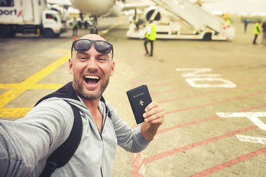 Excited young man traveler taking selfie airplane tarmac holding passport travel document departure airport terminal vacation journey flight boarding tourism holiday exploration transportation