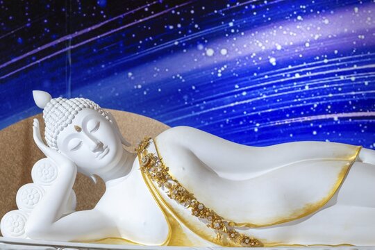 A reclining Buddha, crafted from white material, rests in the classic Parinirvana pose, golden ornaments and floral patterns highlight the fine craftsmanship, whilst the deep blue, cosmic-looking background creates a spiritual, almost ethereal atmosphere. Main chedi (Phra That), Wat Phra That Sorn Kaew, Khao Kho Mountains, Phetchabun Province, Isan, Thailand