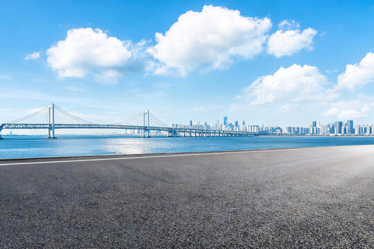 A wide empty asphalt road stretches along a waterfront, with a long suspension bridge and modern city skyline across calm blue water under a bright sky with white clouds.