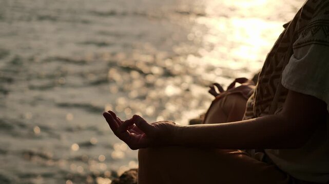 Close-up of a person's hands holding gyana mudra gesture while practicing outdoor yoga meditation by the sea at sunset with warm golden light reflections on the ocean water.