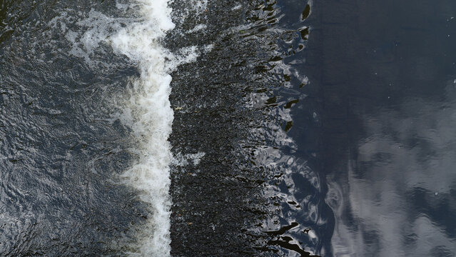 Top down view of flowing water streaming over a small dam or weir threshold creating white foam, splashes, and dark water reflections. Abstract aquatic texture and movement pattern, ideal for nature, 