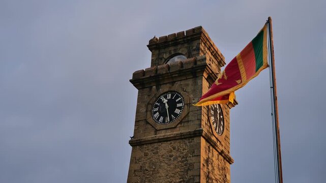 Low-angle view of the historic Galle Fort Clock Tower made of stone with the Sri Lankan national flag waving in the wind against a cloudy sunset sky.