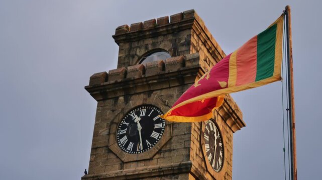 Low-angle view of the historic Galle Fort Clock Tower made of stone with the Sri Lankan national flag waving in the wind against a cloudy sunset sky.