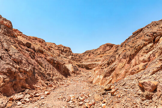 A dry rocky gorge in an arid desert landscape under a clear bright blue sky, showing rugged geological formations.