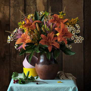 wildflower bouquet with orange daylilies sits in a ceramic vase next to stacked bowls. Floral decoration, botanical arrangement, and rustic still life concept