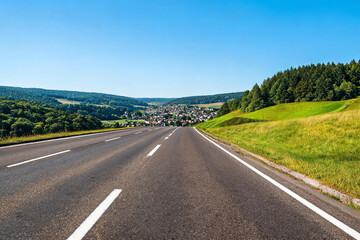 Fototapeta premium A two-lane asphalt highway stretches toward a small town nestled in rolling green hills under a clear blue sky.