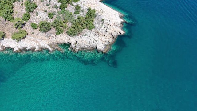 Oblique aerial view moving over a rugged rocky shoreline bordered by transparent turquoise shallows and deep blue sea.
