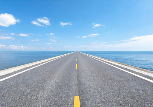 A straight asphalt road stretches across the sea under a clear blue sky with scattered white clouds.