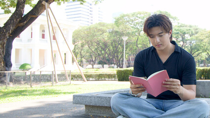 Fototapeta premium Thai Male University Student Reading a Pink Book in a Sunny Park