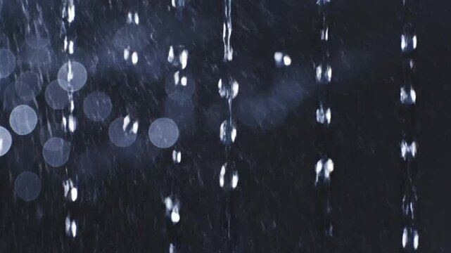 Close-up of falling water droplets creating a shimmering effect against a dark, blurred background with bokeh lights.