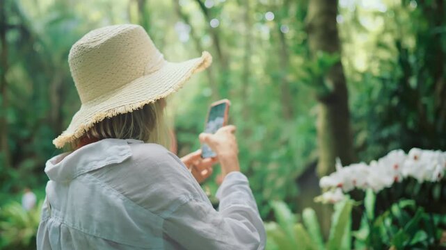 Tourist woman taking mobile photo of beautiful flowers in rainforest, greenhouse . Lady capturing unique beauty of nature and happy impression of tourist trip by smartphone, social media addiction