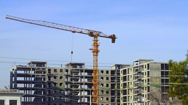 Video of a construction site in which work is underway to erect a multi-storey building using a yellow tower crane. The hook and free berths are lifted, the slinger gives hints to the crane operator