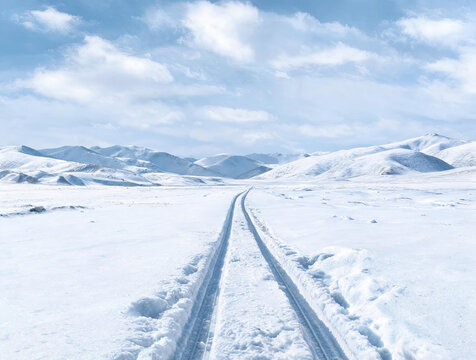 A vast snowy landscape with two parallel tire tracks leading toward distant snow-capped mountains under a bright blue sky with scattered clouds.