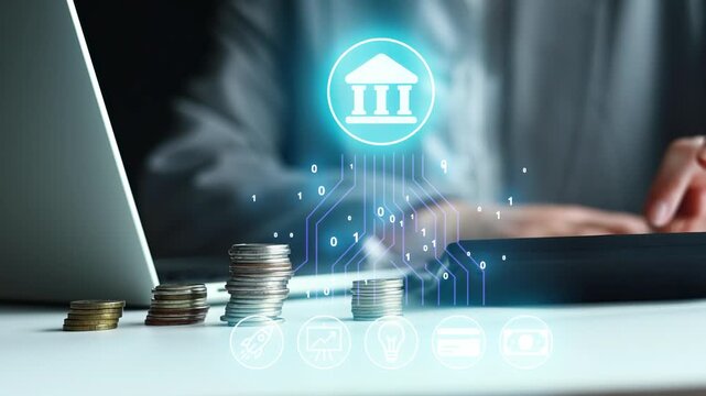 Stacks of coins are placed on a table next to a laptop. A person is using a device, and graphics of banking symbols are displayed above. This scene is in an office setting.