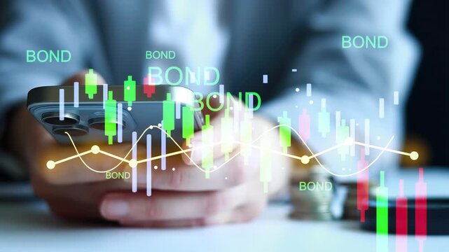A person holds a smartphone showing bond market data with charts and graphs. Documents and coins are visible on the table. This takes place during work hours in an office.