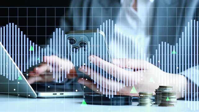 A person is using a smartphone while working on a laptop at a desk. Coins are placed on the desk, and financial graphs show market trends in the background.