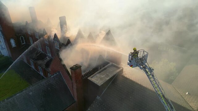 Drone shot of a firefighter on an aerial ladder hosing a burning building as thick smoke billows from the collapsed roof