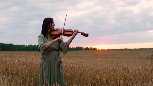 Woman in flowing green dress playing violin in golden wheat field at peaceful sunset, creating serene countryside music under dramatic cloudy sky