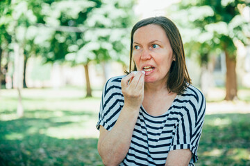 Fototapeta premium Middle-aged woman applying lip balm while sitting in a sunny city park. Self-care, skincare, personal wellness and healthy outdoor lifestyle concept.