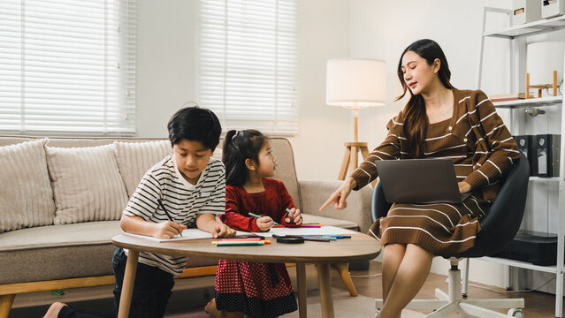 Asian mother working on laptop with two children drawing at home