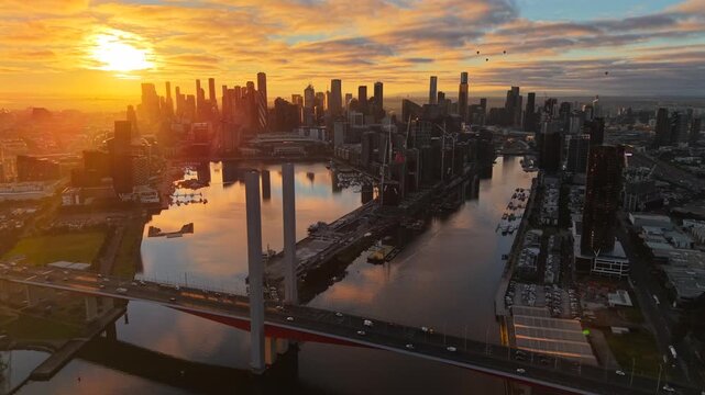 Aerial drone video from behind Bolte Bridge at sunrise, showing Docklands waterways and the full Melbourne CBD skyline under golden morning light