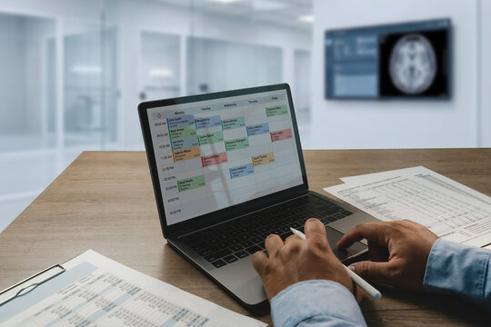 Doctor reviews reports while using laptop displaying a weekly medical appointment schedule supporting healthcare scheduling, clinical administration, and medical workflow planning.