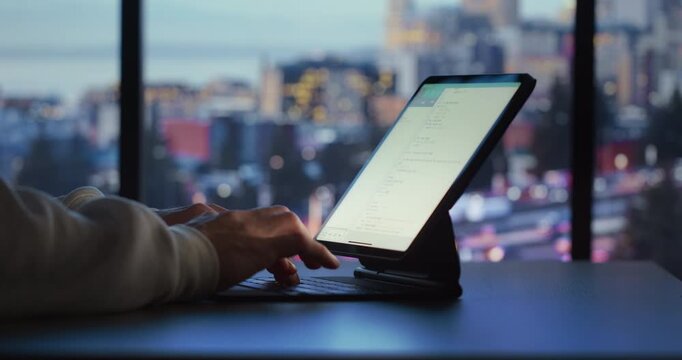 A person types on a tablet with an attached keyboard by a window overlooking a glowing city skyline at night.