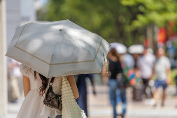 Rear view of a woman carrying a parasol on a city street in dappled light during summer heat with a blurred pedestrian crowd background. © thomasgweber