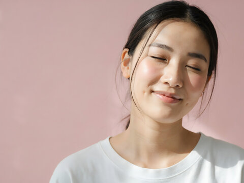 Young asian woman with eyes closed and a gentle smile, posing against a soft pink background, highlighting natural beauty and clear skin