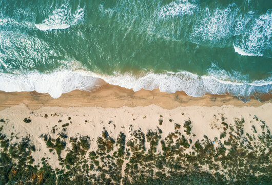 Aerial view shows vibrant green ocean water with white foam waves crashing onto a tan sandy beach.