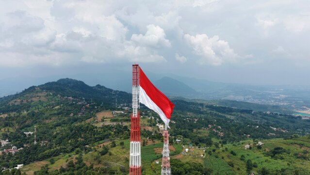 Drone aerial view of the Indonesian flag waving above a telecommunications tower in a mountain landscape.