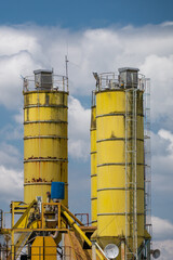 two tall, weathered yellow industrial silos at a concrete batching plant, standing against a vibrant blue sky filled with fluffy white clouds. © Andrey
